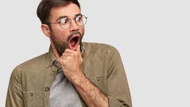 scared anxious male keeps hand near opened mouth looks with very frightened expression aside notices something awful wears round spectacles fashionable shirt poses against white wall 273609 15407